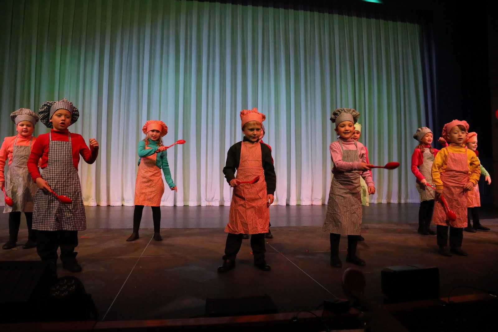 Children in chef's aprons and hats hold large red spoons.