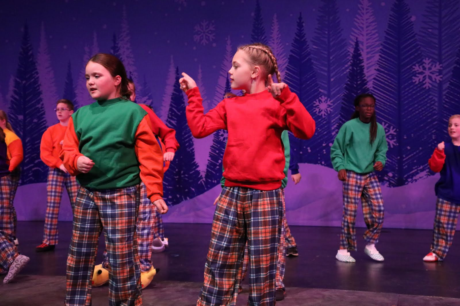 Children in plaid pajamas gesture in front of a Christmas tree drop.